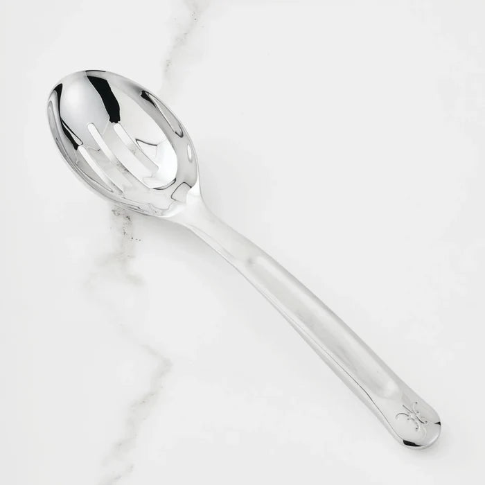 The 10.5-inch Stainless Steel Slotted Serving Spoon from Hestan Provisions® rests on a white marble countertop, its reflective surface gleaming.