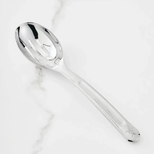 The 10.5-inch Stainless Steel Slotted Serving Spoon from Hestan Provisions® rests on a white marble countertop, its reflective surface gleaming.