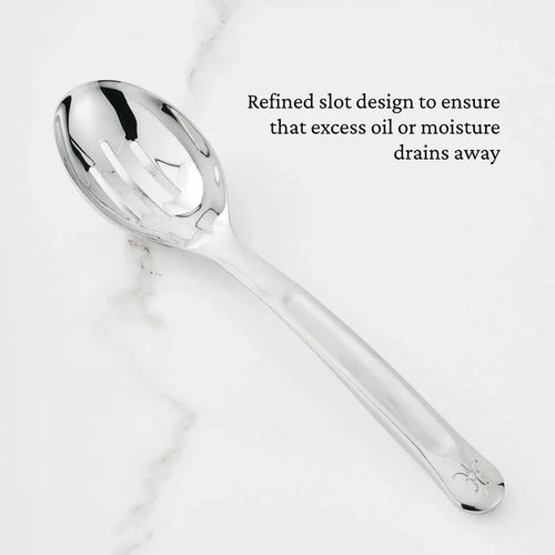A 10.5-inch Stainless Steel Slotted Serving Spoon from Hestan Provisions® rests on a white marble surface. Black text beside the spoon reads, Refined slot design to ensure that excess oil or moisture drains away.