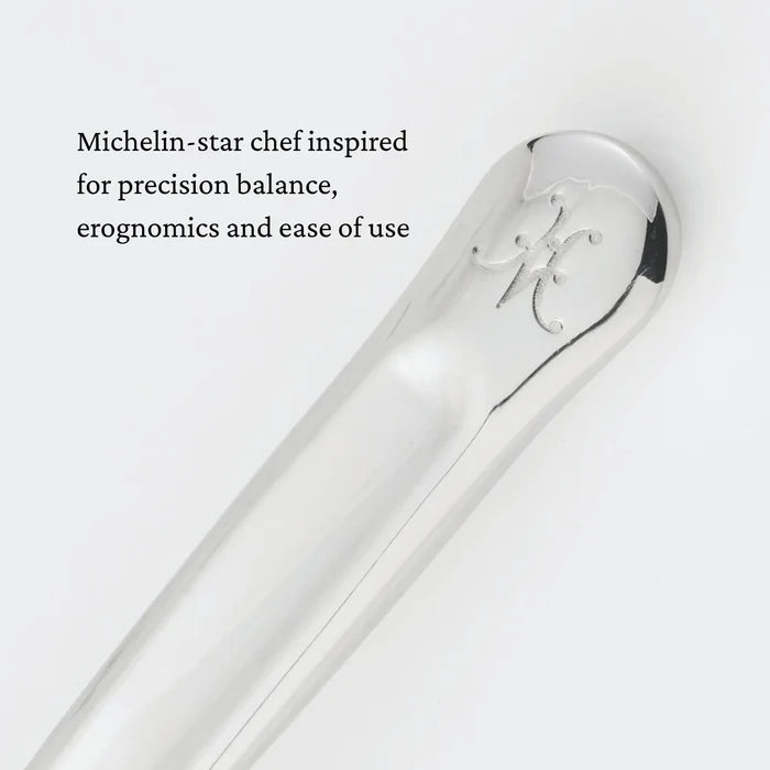 A close-up of a polished metal handle engraved with a decorative letter. Text beside it reads, Michelin-star chef inspired for precision balance, ergonomics, and ease of use. Crafted as part of the Hestan Provisions® Serving Tools collection, the 10.5-inch Stainless Steel Slotted Serving Spoon brings professional-grade quality to your kitchen.