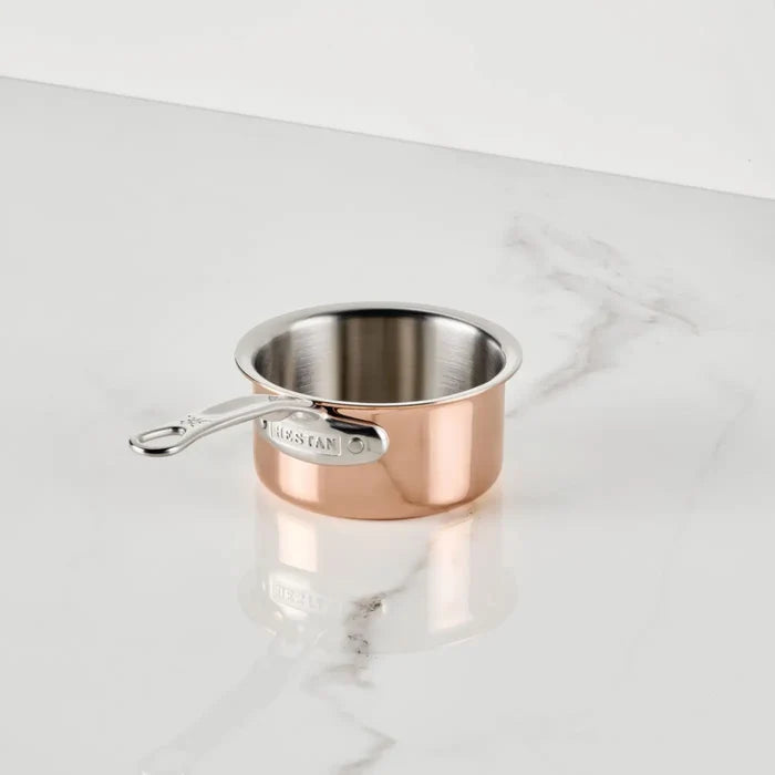The CopperBond Aura™ Polished Copper 0.75-Quart Butter Warmer, crafted as a professional-grade pan, rests on a white marble countertop with subtle gray veining. Its interior features durable stainless steel, and the silver metal handle bears the CopperBond Aura™ brand name embossed on it.