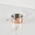 The CopperBond Aura™ Polished Copper 0.75-Quart Butter Warmer, crafted as a professional-grade pan, rests on a white marble countertop with subtle gray veining. Its interior features durable stainless steel, and the silver metal handle bears the CopperBond Aura™ brand name embossed on it.
