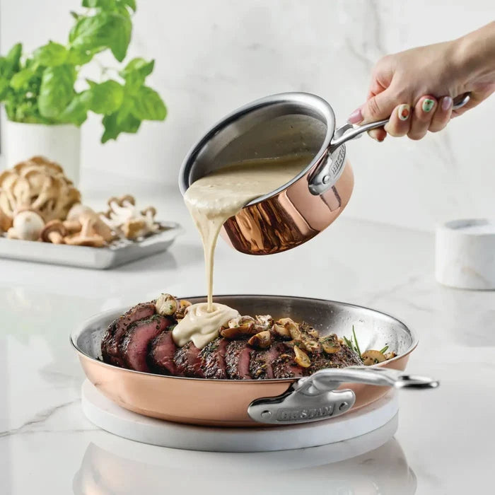 A hand pours creamy sauce from the CopperBond Aura™ Polished Copper 0.75-Quart Butter Warmer onto sliced steak with mushrooms in a professional-grade copper pan. Fresh herbs and mushrooms are arranged on a plate in the background.