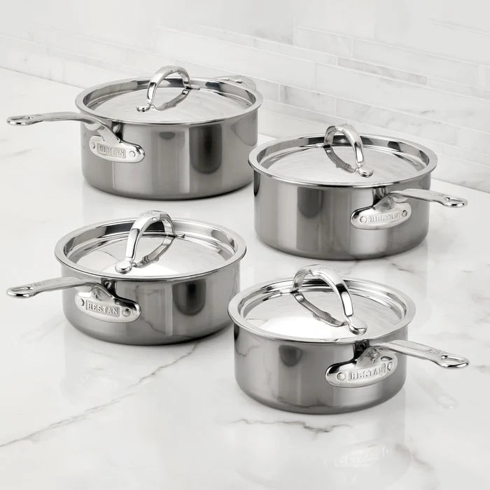 Four NanoBond® Titanium Saucepans with matching lids are arranged on a white marble countertop. The saucepans, featuring advanced molecular titanium cookware technology, come in varying sizes and are equipped with long handles and branded nameplates on the sides. A tiled backsplash appears in the background.