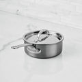 A NanoBond® Titanium Saucepan with a lid rests on a white marble countertop in a modern kitchen. This titanium stainless steel saucepan features a sleek metal handle and polished finish, with the NanoBond® brand name clearly visible on the side.