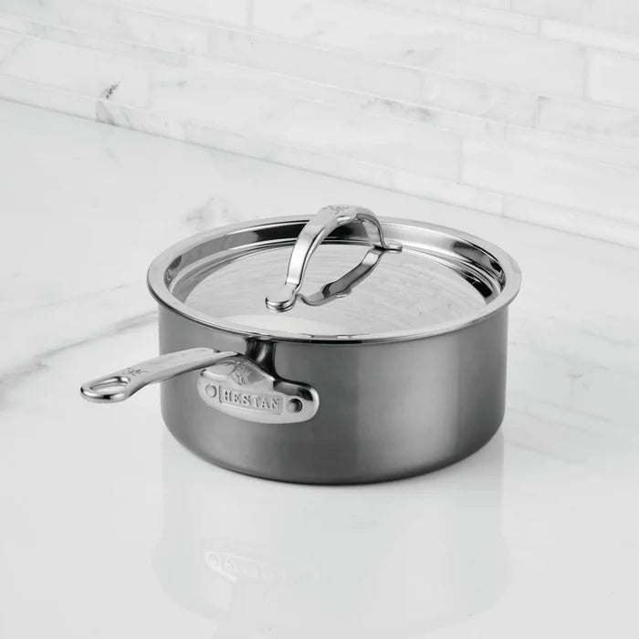 A NanoBond® Titanium Saucepan with a lid rests on a white marble countertop. Crafted from molecular titanium cookware, this stainless steel saucepan features a curved handle on the lid and a long handle labeled NanoBond®. The background displays a clean white tile wall.