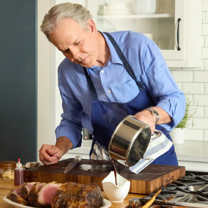 A silver-haired man in a blue shirt and apron pours sauce from a small pot—part of his reliable NanoBond® 10-Piece Titanium Ultimate Cookware Set—into a white gravy boat beside a cutting board with a roast. The kitchen features white tile, cabinetry, and a small potted plant in the background.